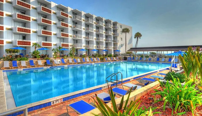 The image shows a bright sunny day at a hotel with a large outdoor swimming pool lined with lounge chairs and shaded by blue umbrellas flanked by a multi-story white building with balconies