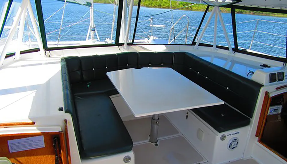 The image shows the interior of a boat with a seating area around a white table overlooking a view of the water through surrounding windows