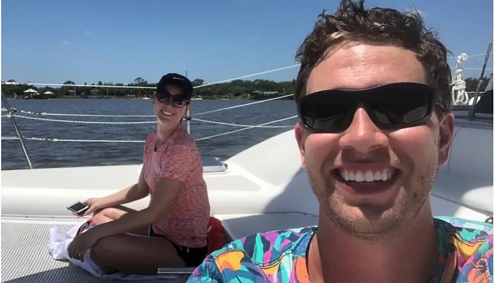 Two people are smiling and enjoying a sunny day on a sailboat taking a selfie with clear skies and calm waters in the background