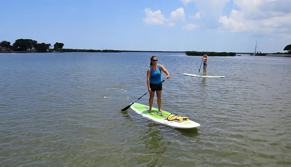 Two people are stand-up paddleboarding on a calm water surface with one in the foreground actively paddling and another standing on her board in the background
