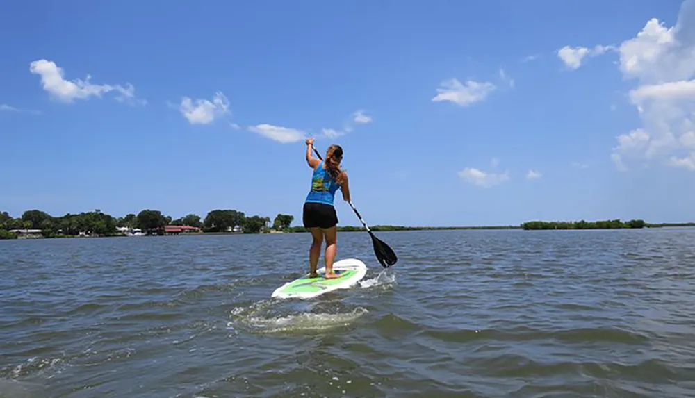 A person is stand-up paddleboarding on a calm body of water under a blue sky dotted with clouds