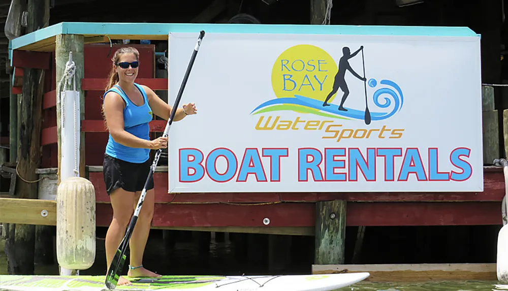 A smiling person is posing with a paddle next to a Rose Bay Water Sports Boat Rentals sign at a water sports rental facility