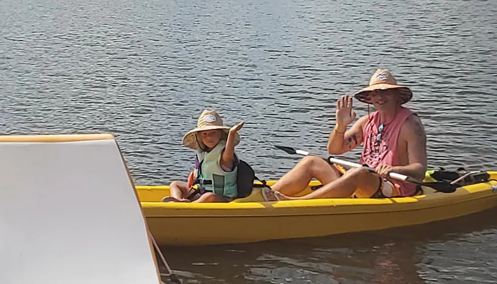 Two individuals are waving and smiling while sitting in a yellow kayak on calm water