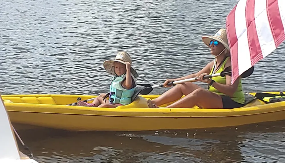Two individuals a child in front sitting and an adult behind rowing are on a yellow kayak on the water with the adult using an oar that has an American flag design