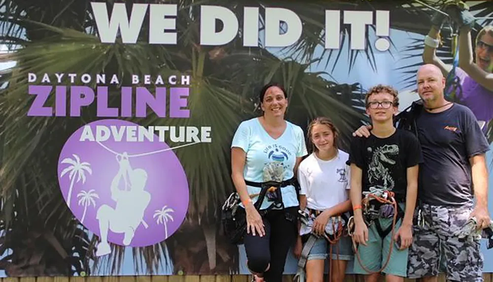 A group of four people possibly a family poses for a photo in front of a Daytona Beach Zipline Adventure backdrop with a celebratory We Did It banner overhead suggesting they have completed a zipline course