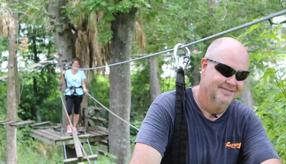 A smiling man in sunglasses is ziplining in a lush green area with another person visible in the background on an elevated platform