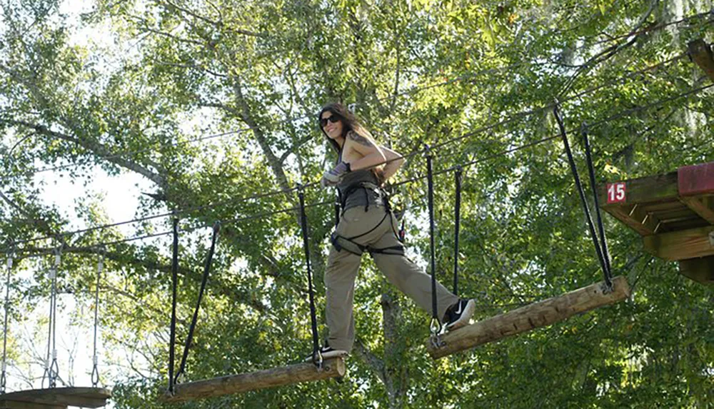 A woman is traversing an outdoor high ropes obstacle course among trees secured with a harness and safety equipment