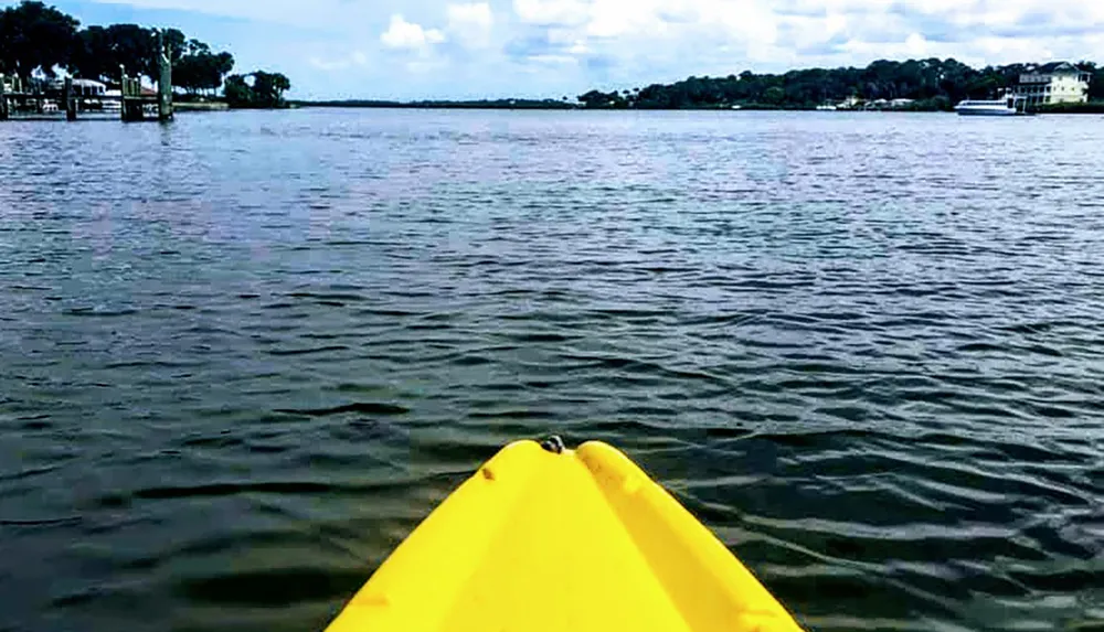 The image shows the point of view from a yellow kayak on a calm body of water with waterfront properties visible in the distance