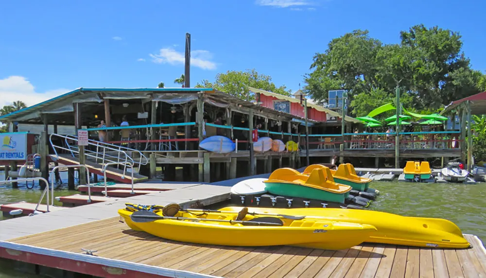The image shows a rustic waterfront establishment with a dock featuring rental kayaks and paddle boats under a clear blue sky