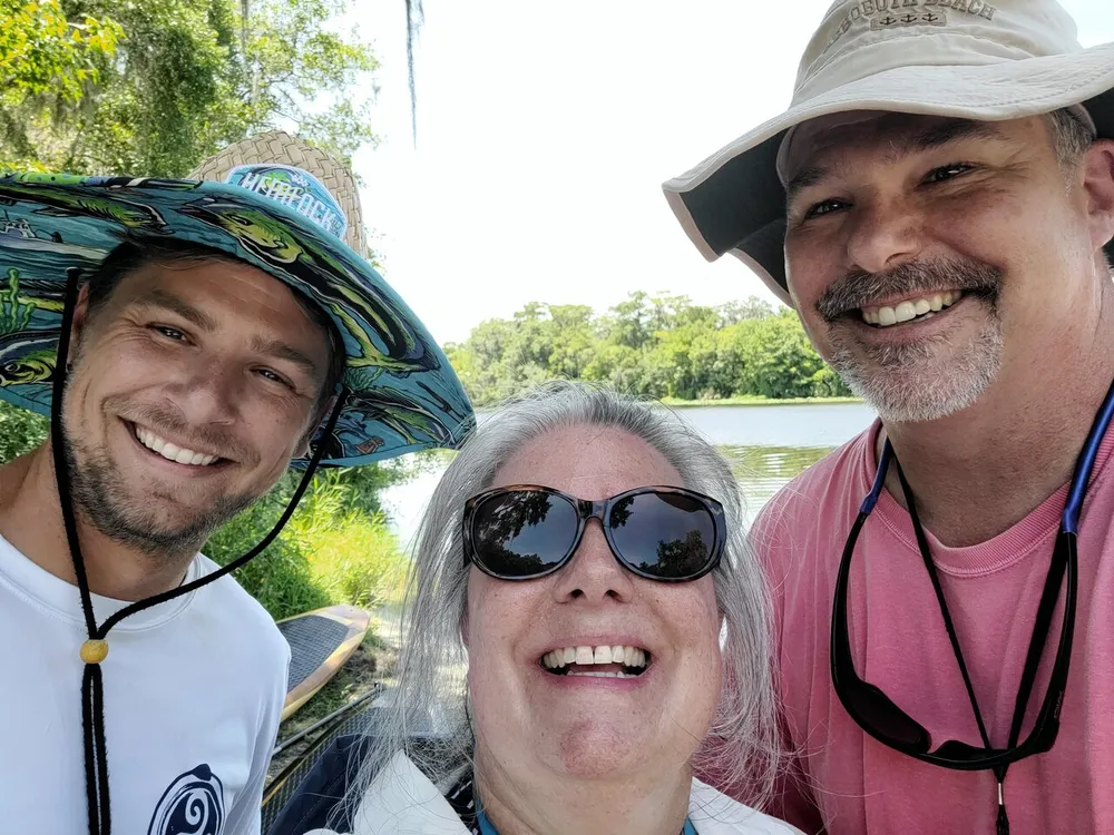 Three people are smiling for a selfie with a natural backdrop with the person in the middle wearing sunglasses and the other two sporting wide-brimmed hats