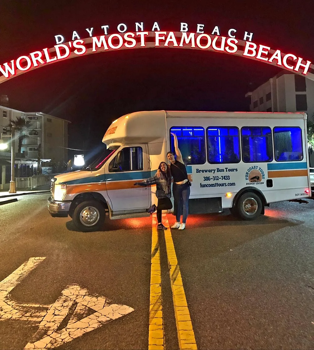 Two individuals pose for a photo in front of a shuttle bus underneath a large arch reading Daytona Beach - Worlds Most Famous Beach at night