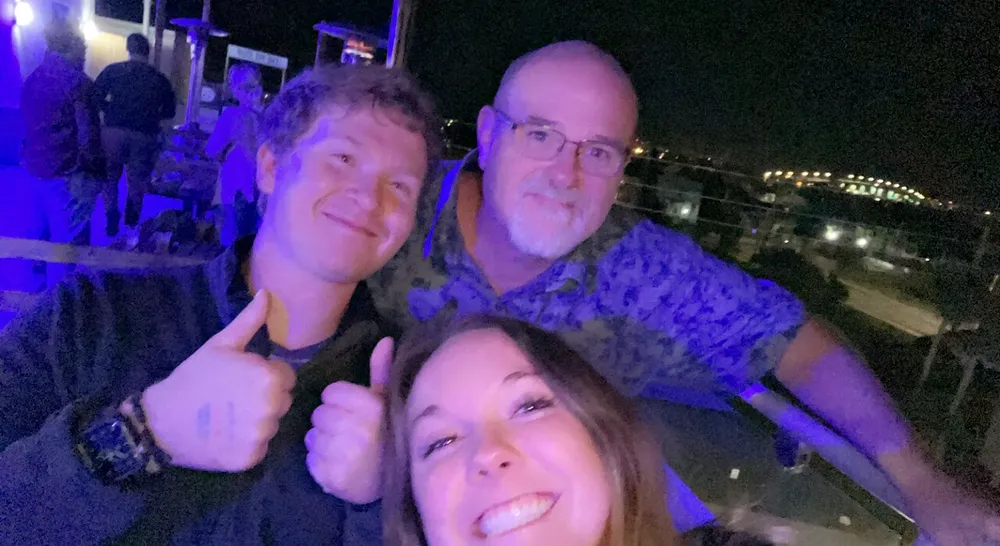 Three people are taking a cheerful selfie together at night with ambient lights in the background suggesting they might be at an outdoor event or venue