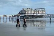 Two people ride Segways on a wet beach with a large pier structure in the background.