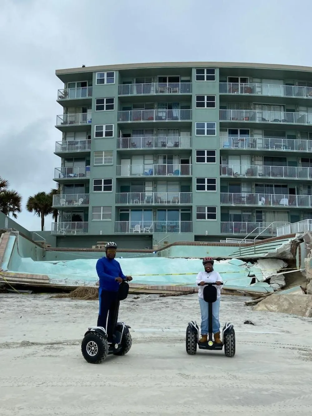 Two individuals are riding Segways on a beach with a damaged building in the background