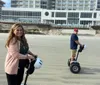 Two people ride Segways on a wet beach with a large pier structure in the background