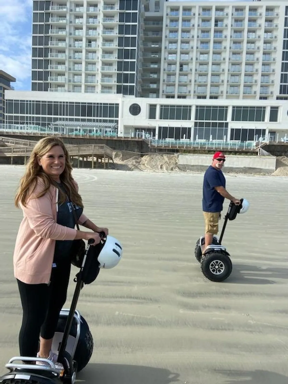 Two people are enjoying a ride on Segways along a sandy beach with a large hotel building in the background