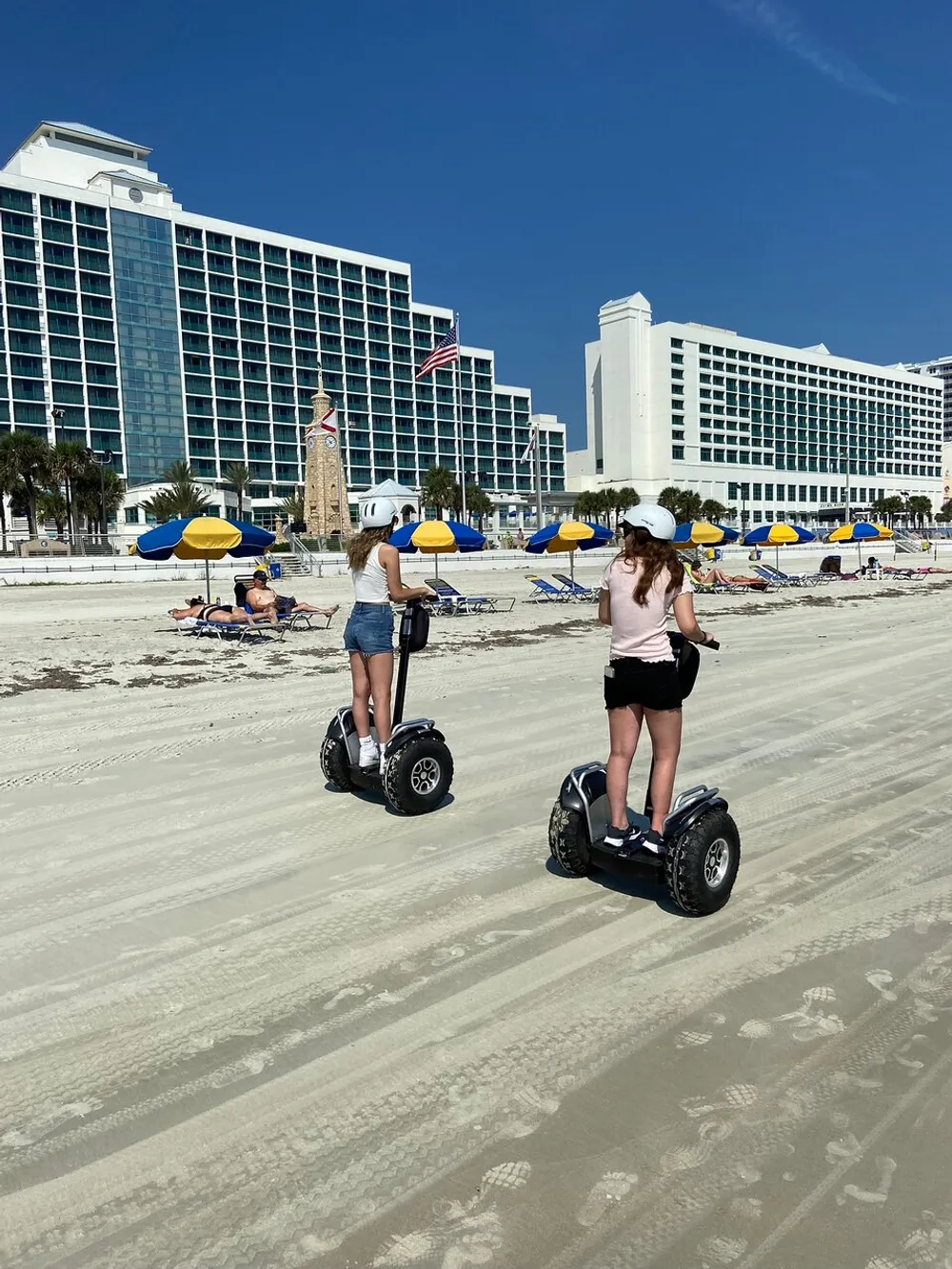 Two people wearing helmets are riding Segways on a sunny beach with umbrellas sun loungers and a large hotel complex in the background