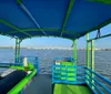 The image shows the view from the rear of a colorful boat with blue and green benches looking towards a river with a bridge in the distance under a clear blue sky