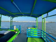 The image shows the view from the rear of a colorful boat with blue and green benches, looking towards a river with a bridge in the distance under a clear blue sky.