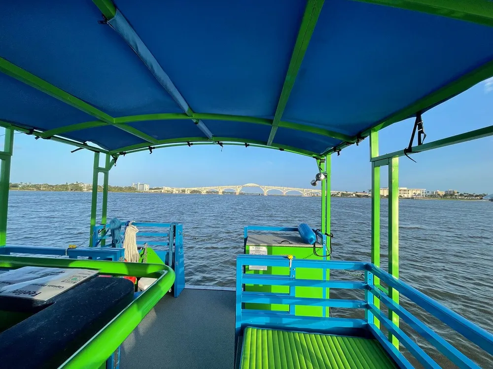 The image shows the view from the rear of a colorful boat with blue and green benches looking towards a river with a bridge in the distance under a clear blue sky