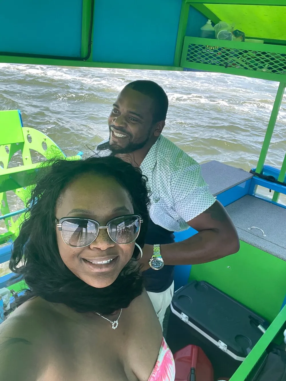 A woman is taking a selfie with a smiling man standing behind her on a boat with vibrant green trim and choppy water in the background