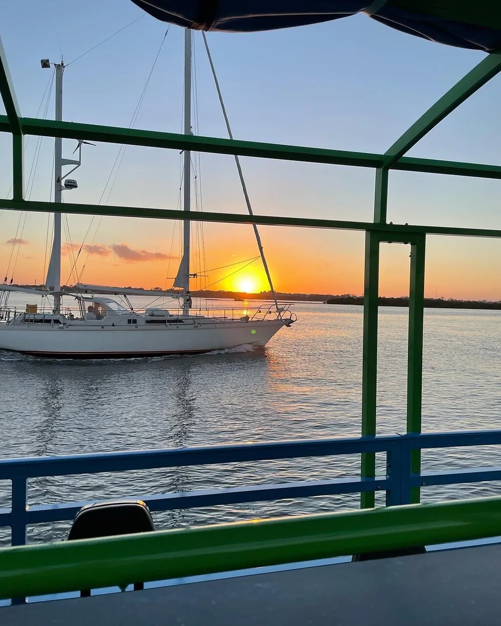 A serene sunset with the sun descending behind a sailboat viewed through the vibrant green structure of a waterfront railing