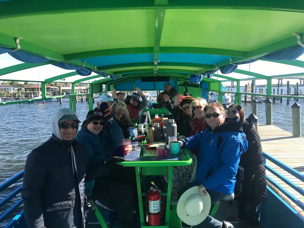 A group of cheerful people dressed in warm clothing are seated at a covered green table aboard a boat waving and enjoying their time together