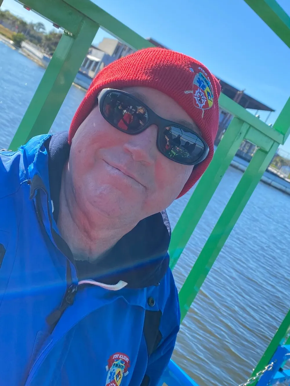 A person in a blue jacket and red beanie is smiling while wearing sunglasses near a green structure with a water backdrop