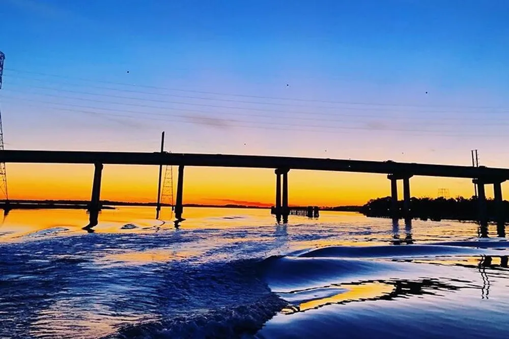 The image depicts a bridge silhouetted against a vibrant sunset sky with reflections on the water below