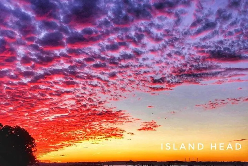 The image shows a vivid sunset with a sky filled with textured clouds that are painted in shades of purple and red with a silhouette of a treeline and the words ISLAND HEAD displayed at the bottom