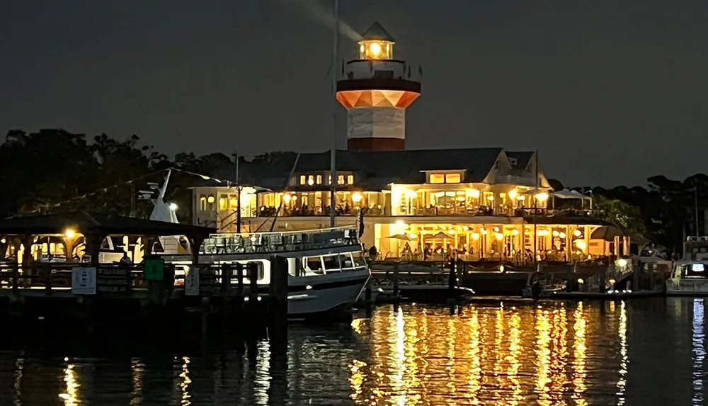 The image shows a lively harbor scene at night featuring a warmly lit multi-story building with a lighthouse tower a dock with boats and reflections on the water