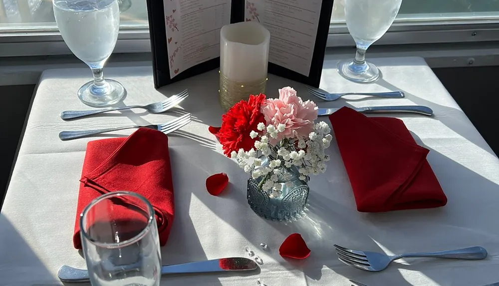 The image shows a romantic table setting with red and pink flowers candles red napkins and scattered rose petals suggesting a special dining occasion