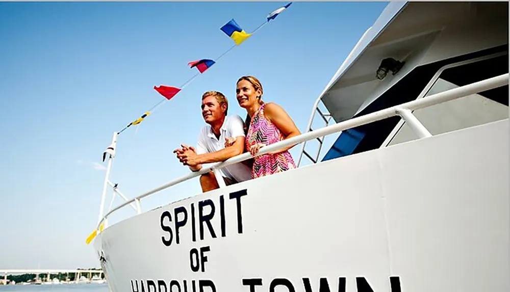 A man and a woman are standing beside the railing of a boat named SPIRIT OF HARROW TOWN adorned with colorful flags looking out towards the horizon