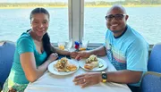 A smiling couple is enjoying a meal together on a boat with a scenic water view in the background.