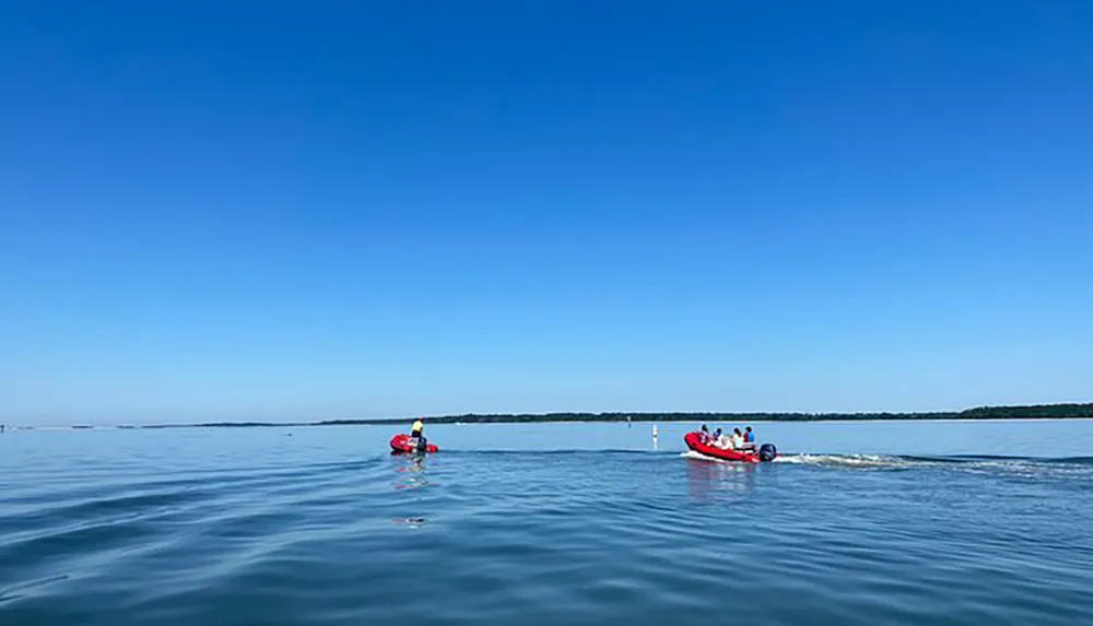 The image shows a serene expanse of blue water under clear skies with a person kayaking in the foreground and a group on an inflatable boat towed by a motorboat in the background