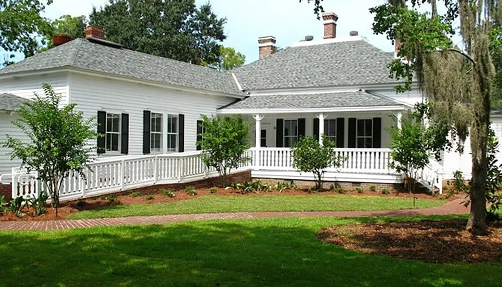 The image shows a single-story white house with black shutters a wraparound porch and a well-manicured lawn with young trees and a brick pathway
