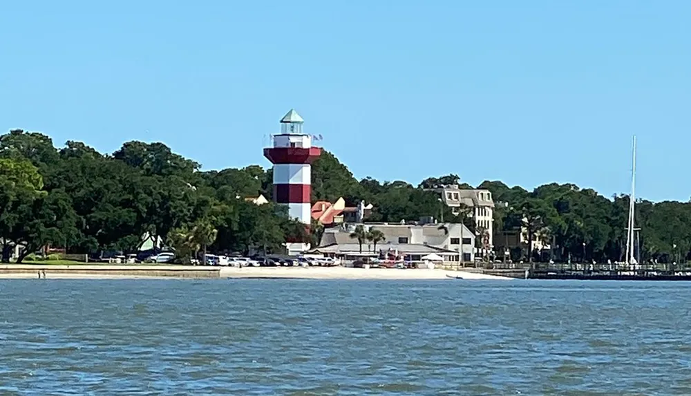 The image shows a picturesque seaside setting featuring a distinctive red and white striped lighthouse near a calm waterfront with parked cars and lush greenery in the background