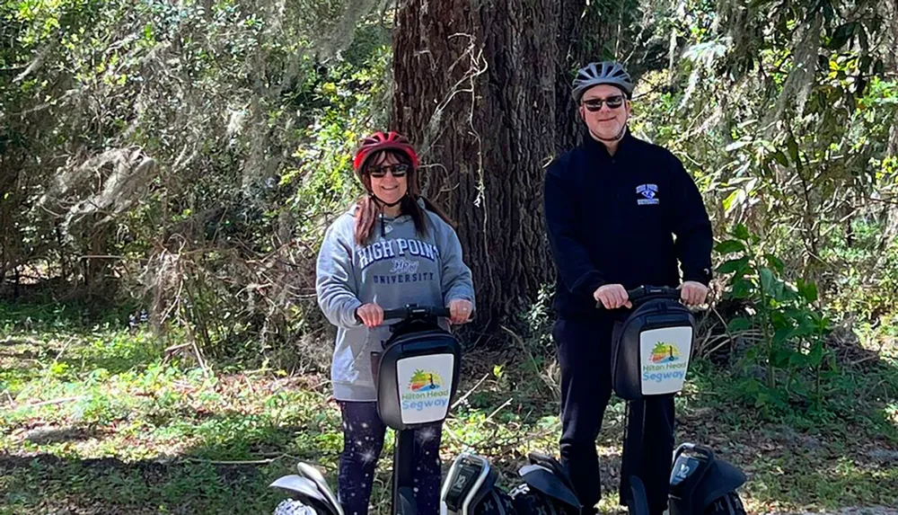 Two individuals wearing helmets are standing with Segways in a wooded area appearing ready for a guided tour or adventure
