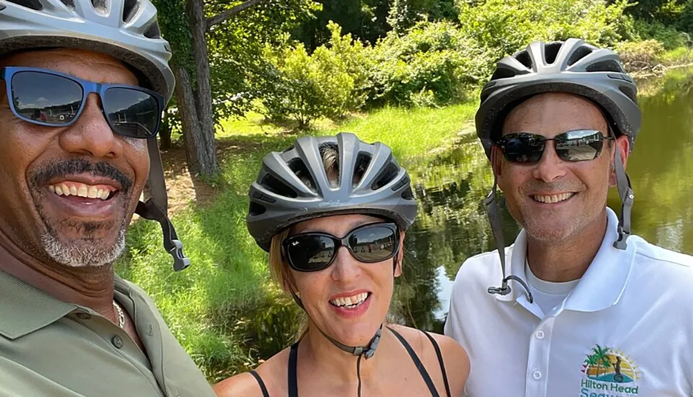 Three smiling cyclists wearing helmets and sunglasses are posing for a photo outdoors with greenery and a water body in the background