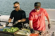 Two chefs are grilling an assortment of fresh vegetables and meats on a boat.