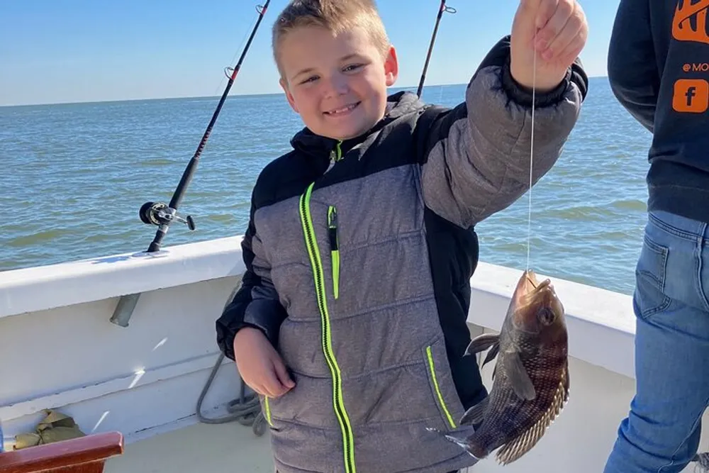 A smiling child on a boat is proudly displaying a fish caught during a fishing trip while a fishing rod rests in the background against the railing of the boat