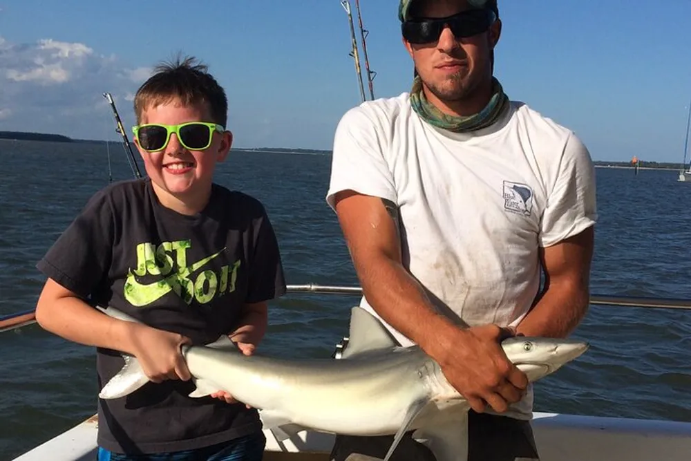 A young person in green sunglasses and an adult are on a boat holding a shark they have caught