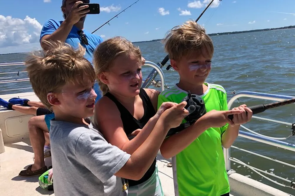 Three children appear focused and excited as they hold a fishing rod together on a boat with a man in the background capturing the moment with a smartphone camera