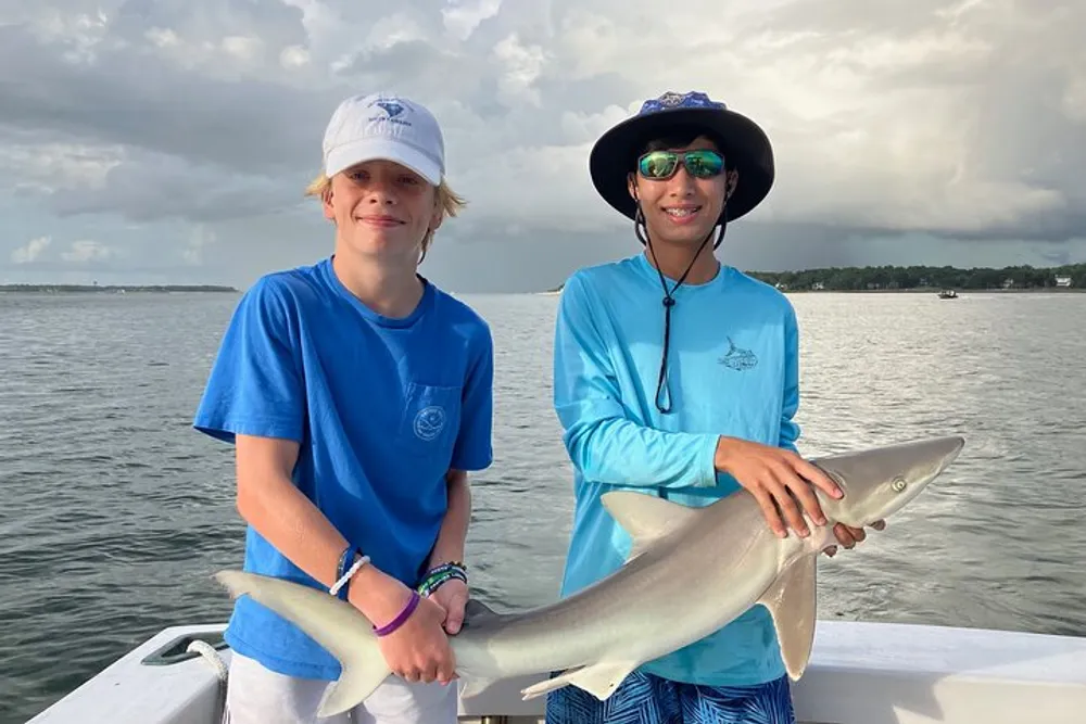 Two smiling young fishermen are holding a shark aboard a boat with cloudy skies and calm waters in the background