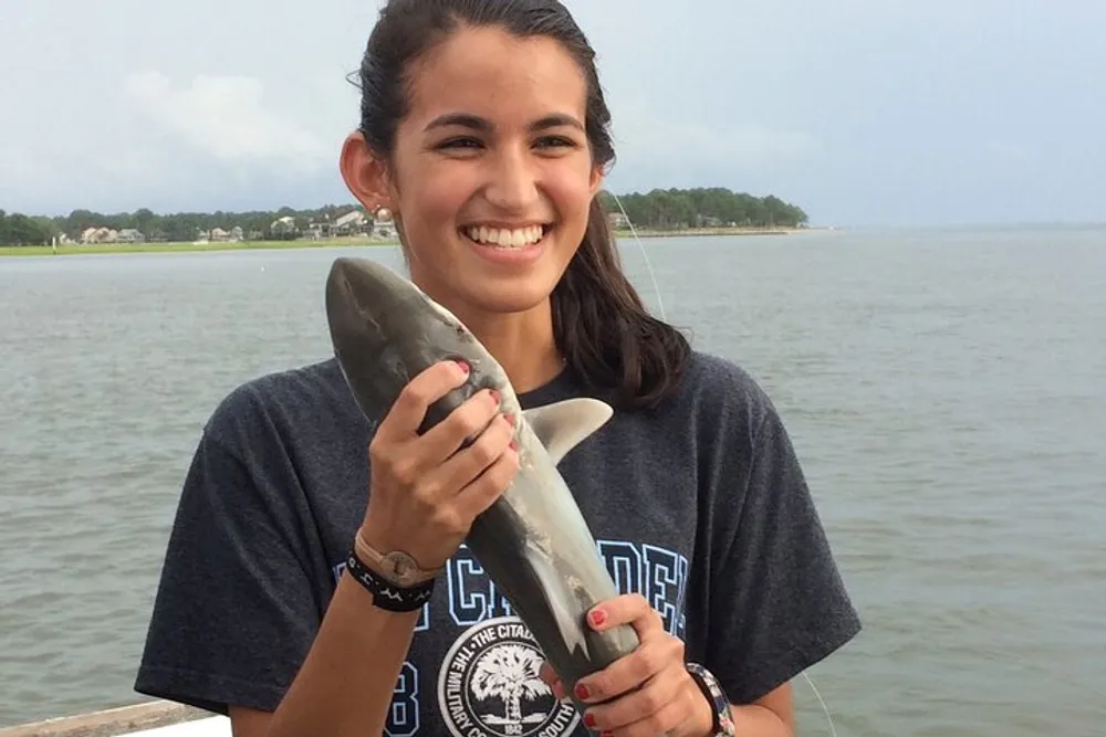 A smiling young woman is holding a small shark by a body of water