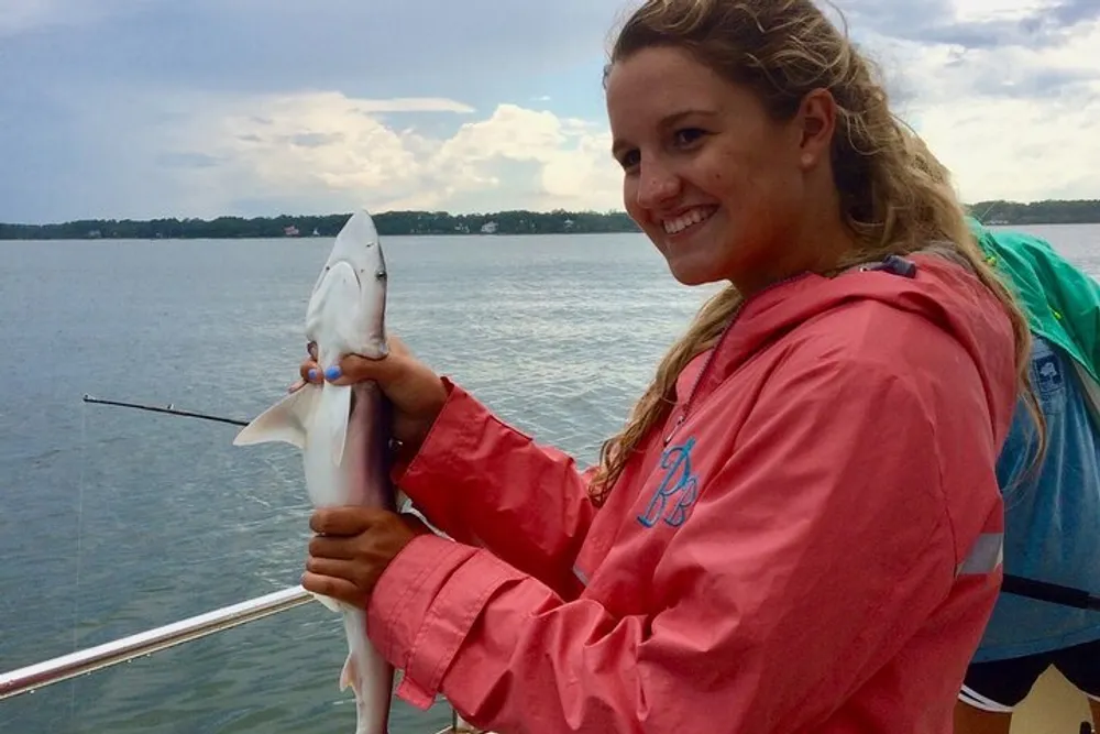A smiling person is holding a small shark on a boat with a body of water and overcast sky in the background