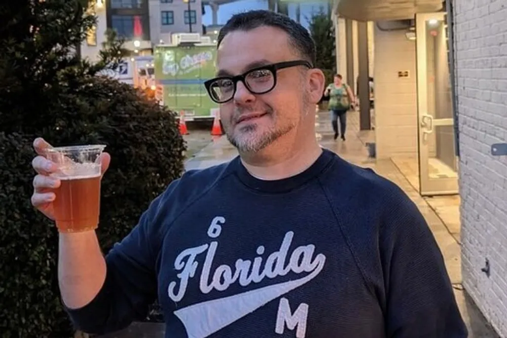 A man in glasses and a Florida sweatshirt is smiling while holding a cup of beer outdoors