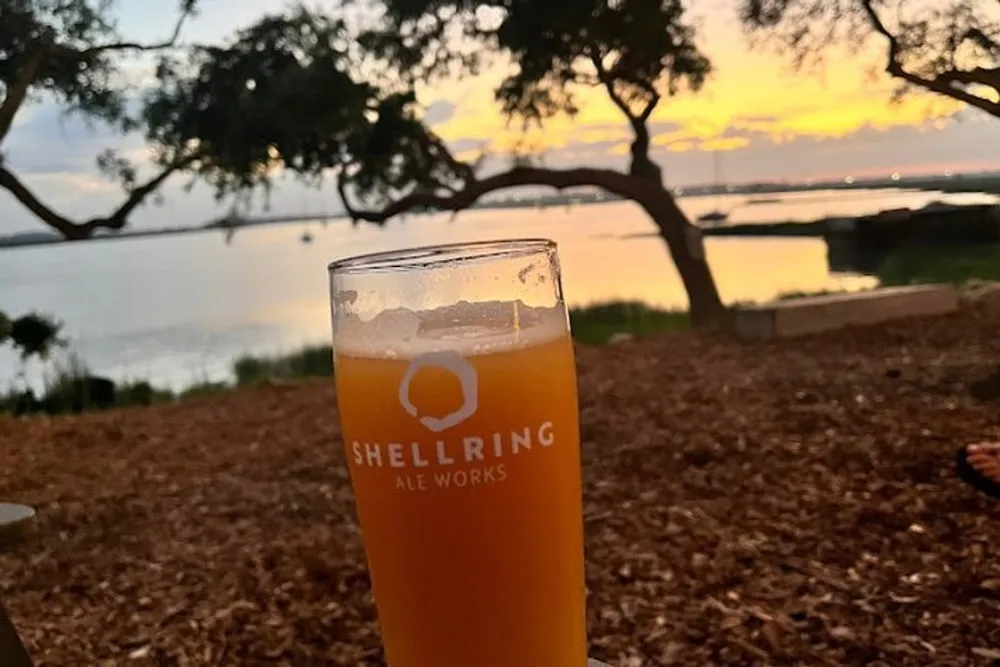A glass of beer rests on a table with a backdrop of a beautiful sunset over a calm waterfront