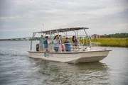 A group of people are enjoying a boat ride on a calm river with lush greenery on one side and a cloudy sky above.