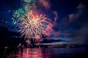A vibrant fireworks display lights up the night sky above a body of water, reflecting colorful bursts on its surface.